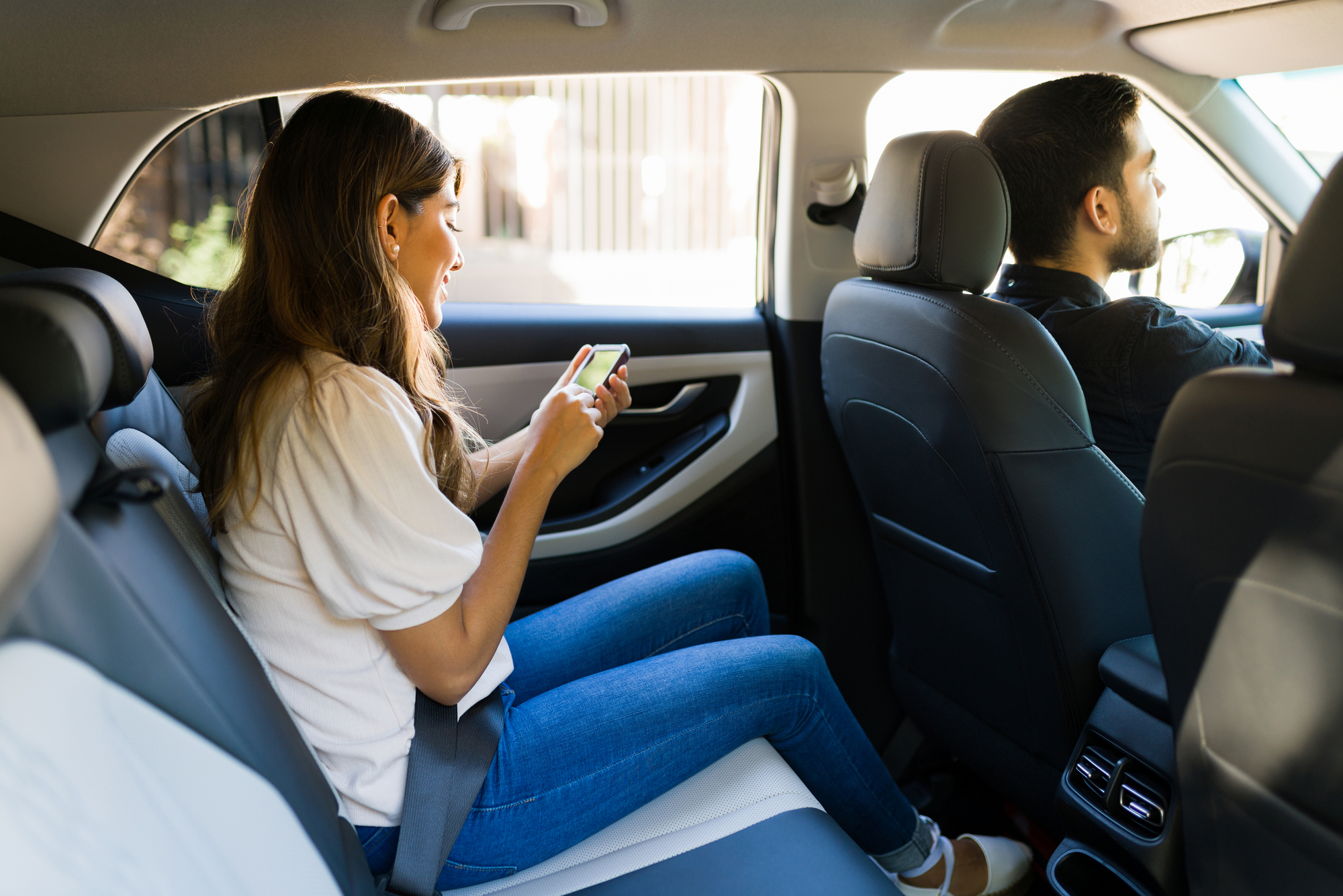 A woman in the back seat of a car looks at her smartphone while a man drives.
